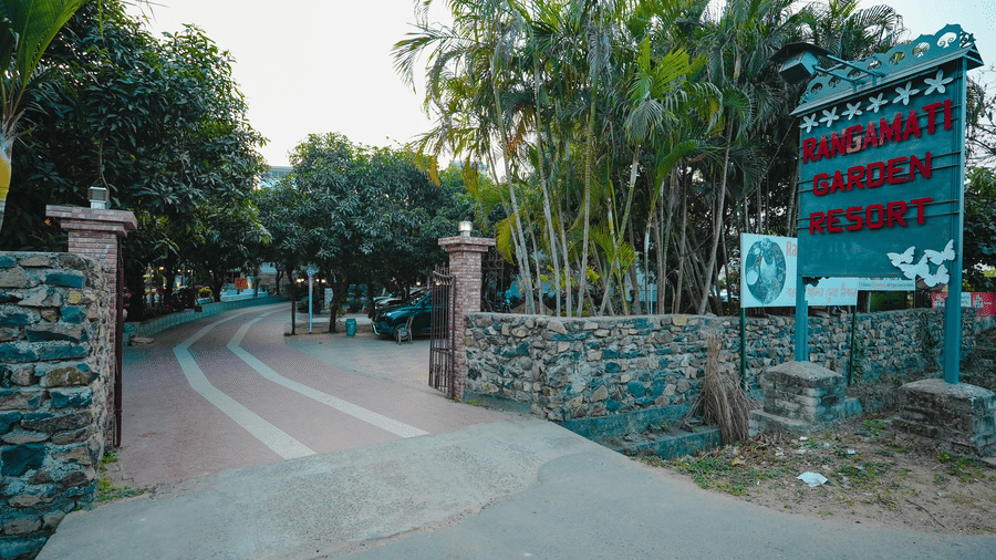 An entrance gateway to a resort featuring a stone wall, paved driveway, lush trees, and a green signpost.
