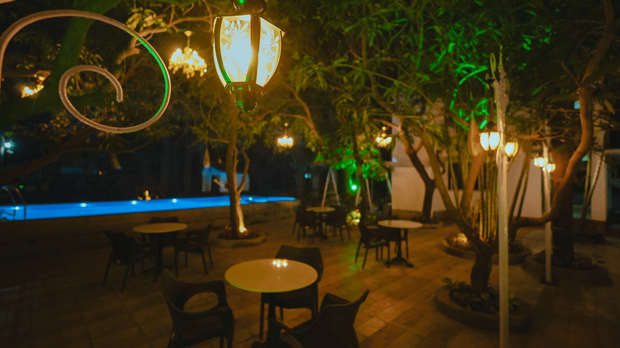 An outdoor restaurant seating area illuminated by warm glowing lamps next to a swimming pool at night.