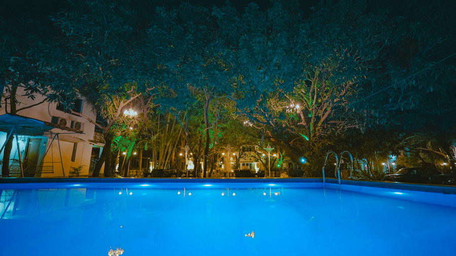 A clear blue swimming pool brightly lit up at night, surrounded by dark silhouettes of trees.