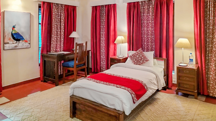 Spacious heritage bedroom with a wooden double bed soft lighting and rich red curtains creating a warm and restful ambience at Neemrana’s Glasshouse on The Ganges with traditional furnishings high ceilings and a calm indoor atmosphere ideal for relaxation