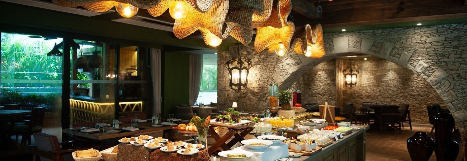 A lavish breakfast buffet spread at Mila Restaurant at The Evren, Vagator, featuring a variety of dishes on a large central island. The restaurant has a rustic yet modern ambiance with woven light fixtures, a stone archway, and a view of the outdoors.