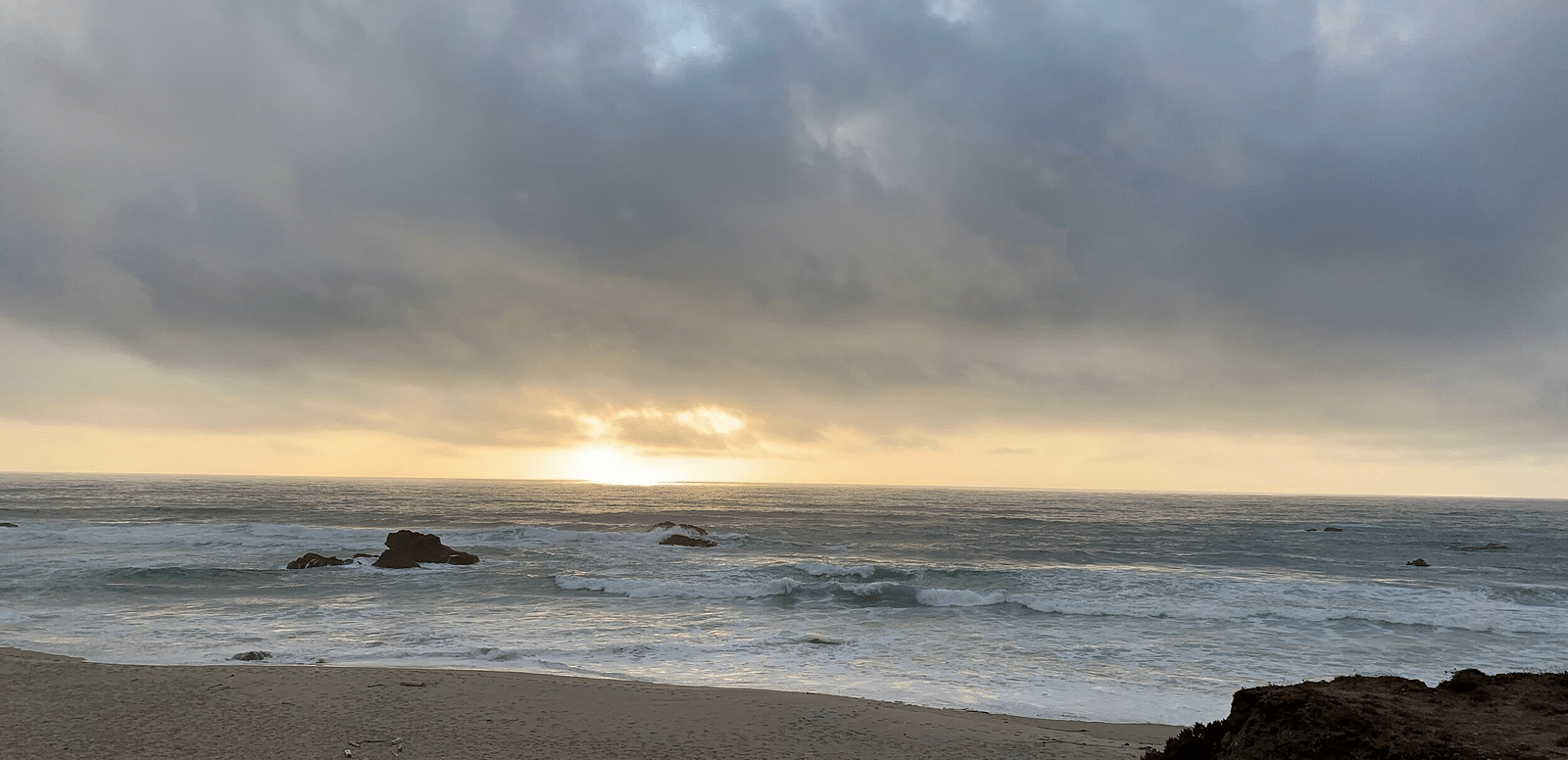 A beach scene with a cloudy sky above the ocean, where the sun is breaking through the clouds on the horizon.