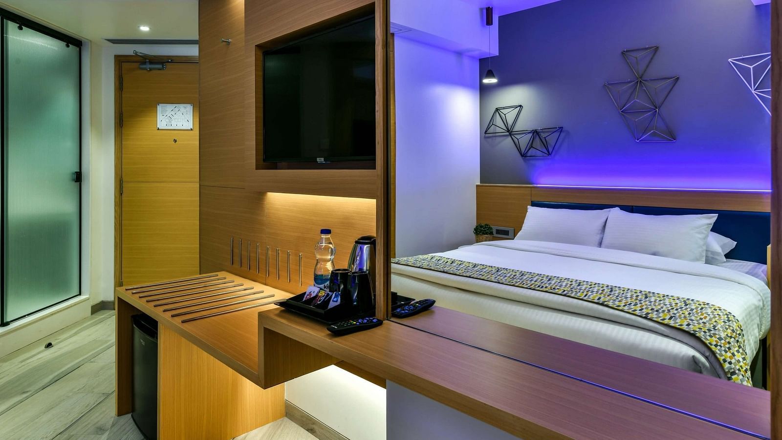 A two-image collage showcasing a spacious bed in one frame and a wall-mounted TV alongside tinted glass sliding doors leading to the washroom in the other, at Indie Stays, BKC Kalina.