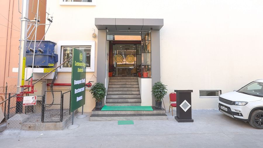 A view of the Lemonridge Hotels KPHB property entrance, showcasing a signboard that displays 'Lemonridge Hotels' at the top and 'Banquet Halls, Restaurant, Rooms' beneath it. Near to it is a transformer unit and a building near it. In the other side of the