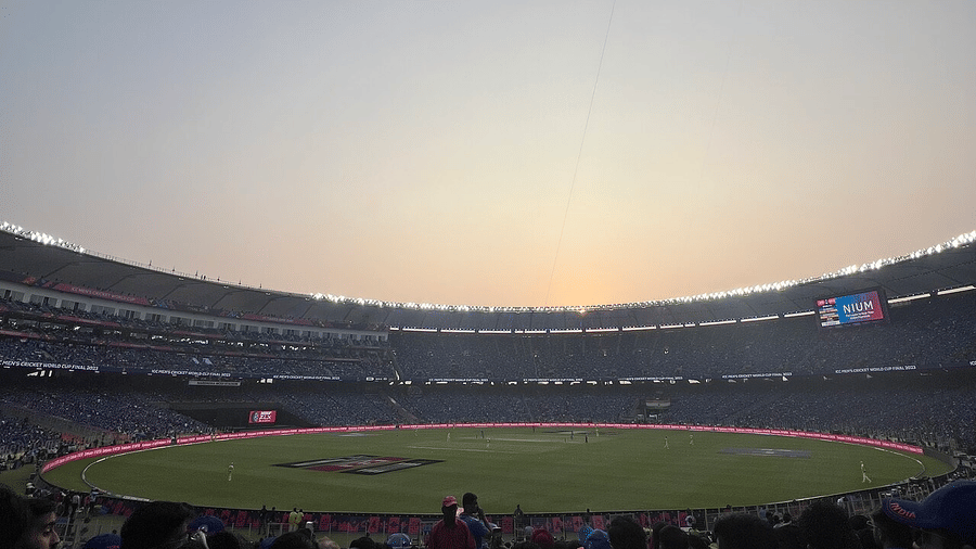 Sunset panorama of cricket stadium, sky glowing orange above the oval as crowd silhouettes ring the boundary.