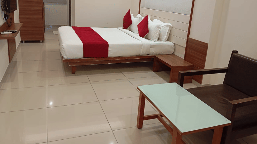 A Deluxe Room at Hotel Ashray Inn Express featuring a large bed, bedside tables, seating area with chairs and center table, wardrobe, and tiled flooring.