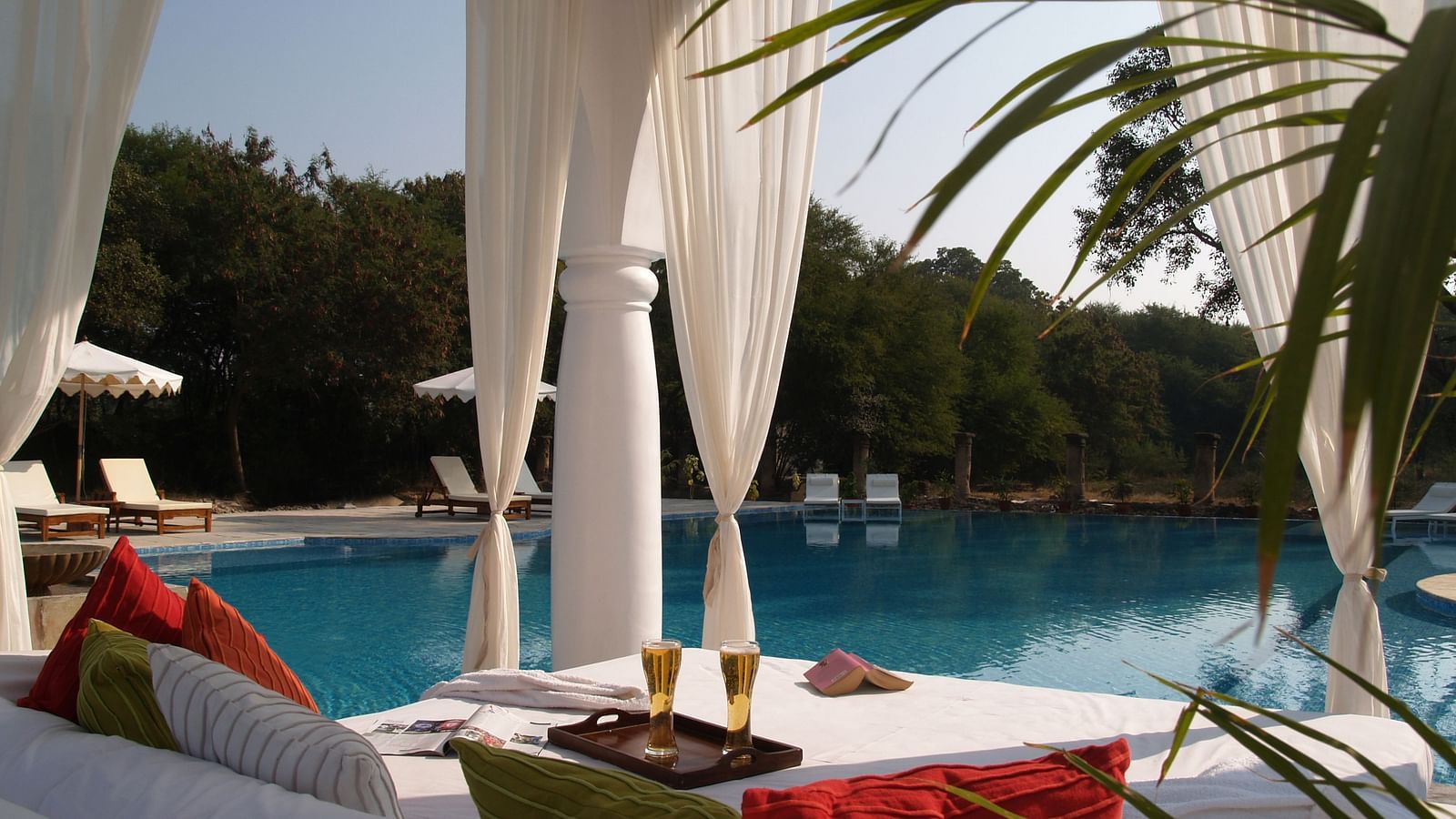 A poolside cabana with white drapes frames a view of a swimming pool, lounge chairs, and trees, with colorful pillows and drinks on a table in the foreground- Shahpura Bagh.