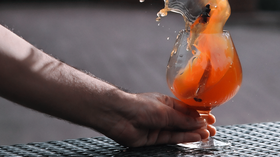 Action shot of a hand holding a glass of orange-coloured drink with ice, as the liquid splashes dramatically out of the glass, set against an outdoor background with greenery - Ananta Spa and Resort, Pushkar.
