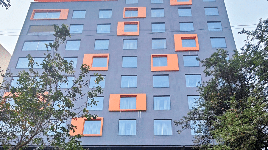A modern, multi-story hotel building with a dark grey facade and bright orange window accents. The hotel has a covered parking area in front, and there are trees and shrubs surrounding the building.