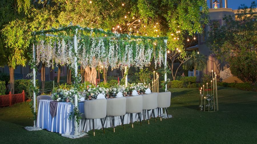An outdoor evening event with a long dining table set for a meal, adorned in a white tablecloth, surrounded by nature and illuminated by overhead string lights.