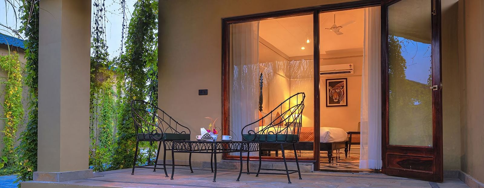 A ground-level view of Superior Cottage's patio featuring a small table and two wrought-iron chairs, with the interior of the room visible through open glass doors - Singhvilas, Ranthambore