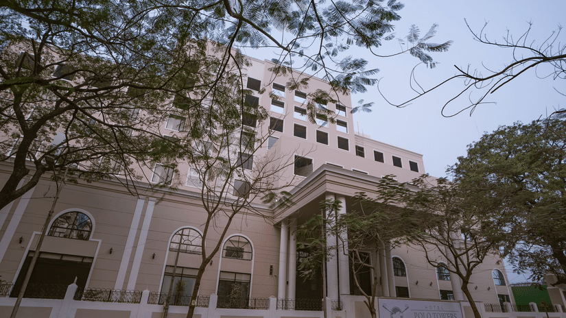 Exterior facade of Polo Hotel, Agartala showing a multi-storey cream building with arched windows, a portico and trees framing the foreground.