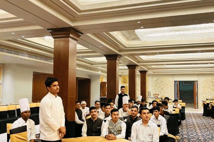 People gather inside a hall at Clarks Group of Hotels. Tables and chairs are arranged across the room for an event.