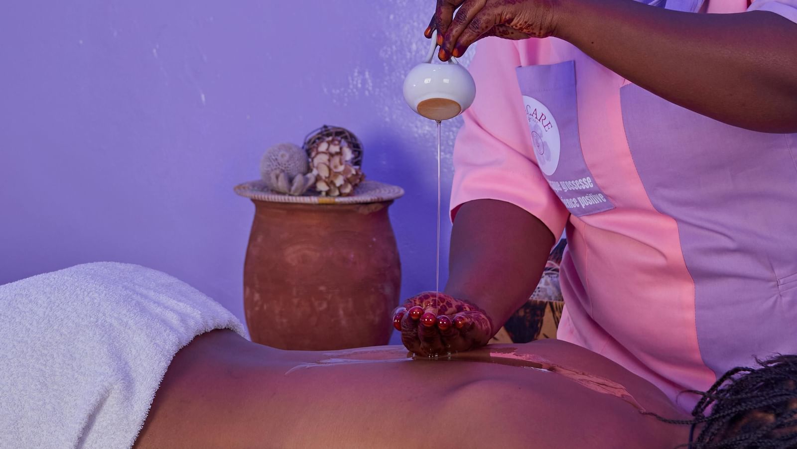  A spa therapist, wearing a headscarf, performs a hot oil massage on a client's back, pouring oil from a small white vessel.