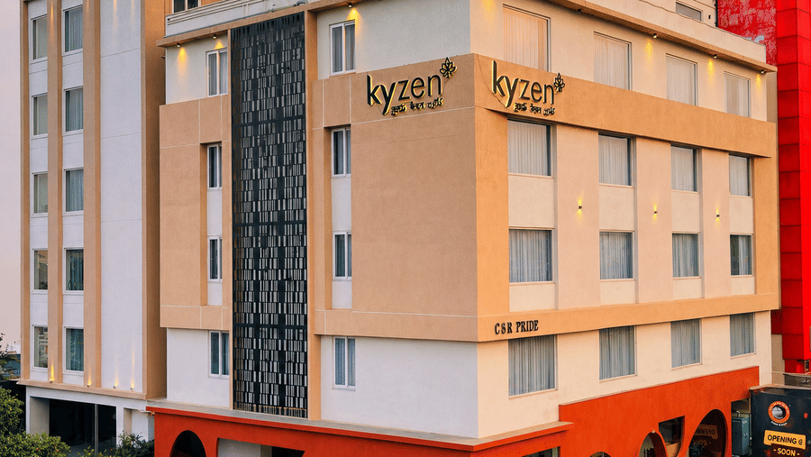 Street level exterior view of Hotel Kyzen HiTech City featuring a multi-storey building with orange base grey upper floors hotel branding and surrounding commercial buildings visible.