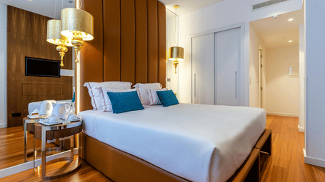 A smart, well-appointed hotel bedroom features a plush bed, warm wooden flooring, and elegant golden bedside lamps.