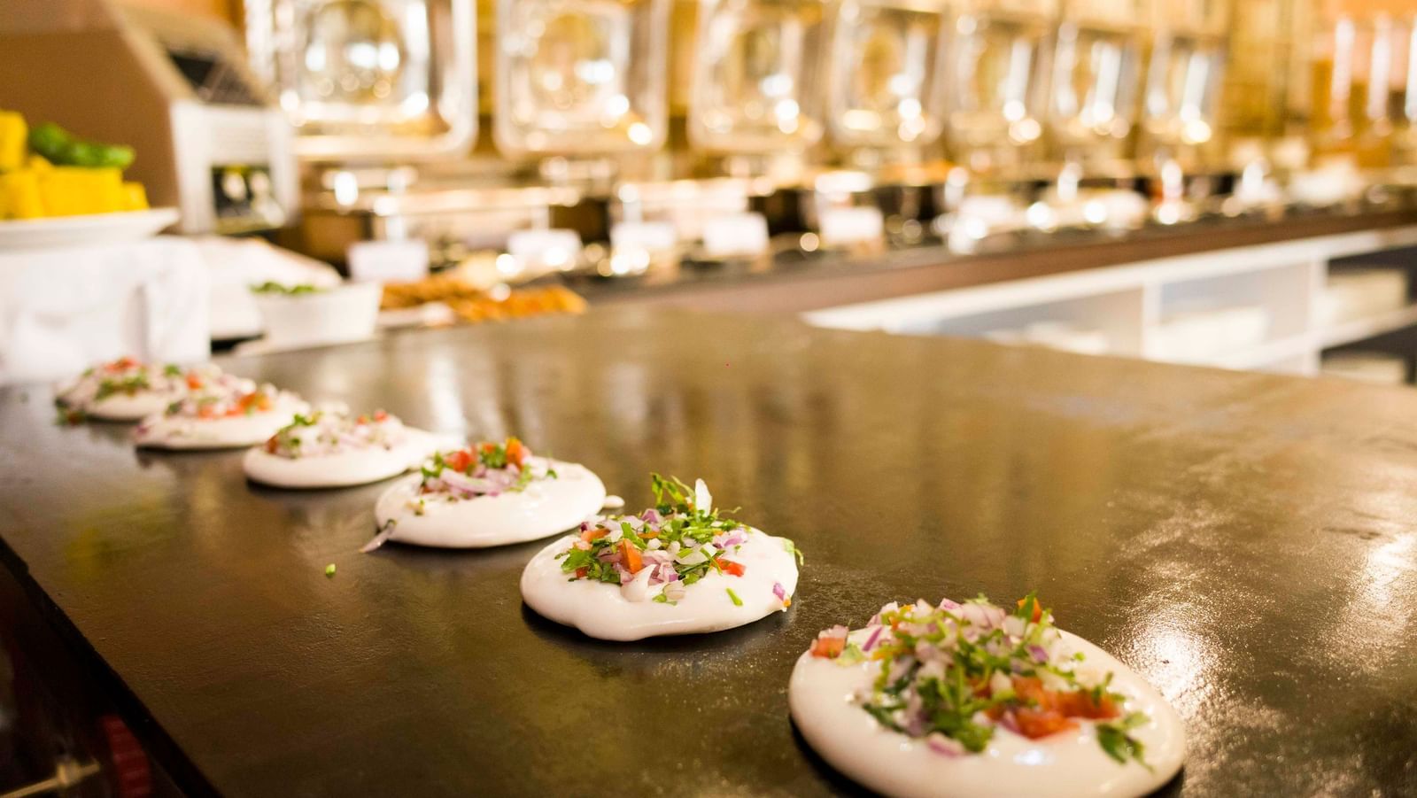 A close-up of small appetizers with colorful toppings on white plates, part of a buffet setup | Daksh The Madhuvan Suites