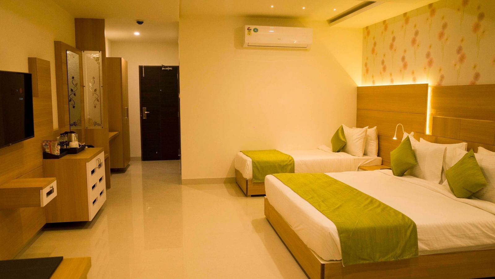 A hotel bedroom with warm lighting, a large bed, a single bed, with low headboards and a wall-mounted television | Daksh Pride Room | Daksh The Madhuvan Suites