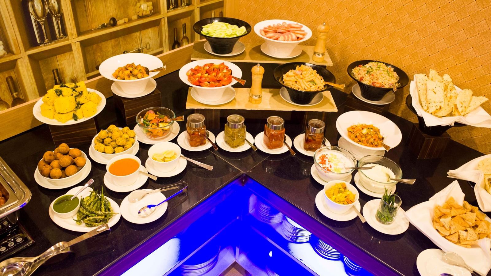 A lavish buffet spread with a variety of appetizers, salads, and condiments, featuring a blue-lit counter element | Daksh The Madhuvan Suites