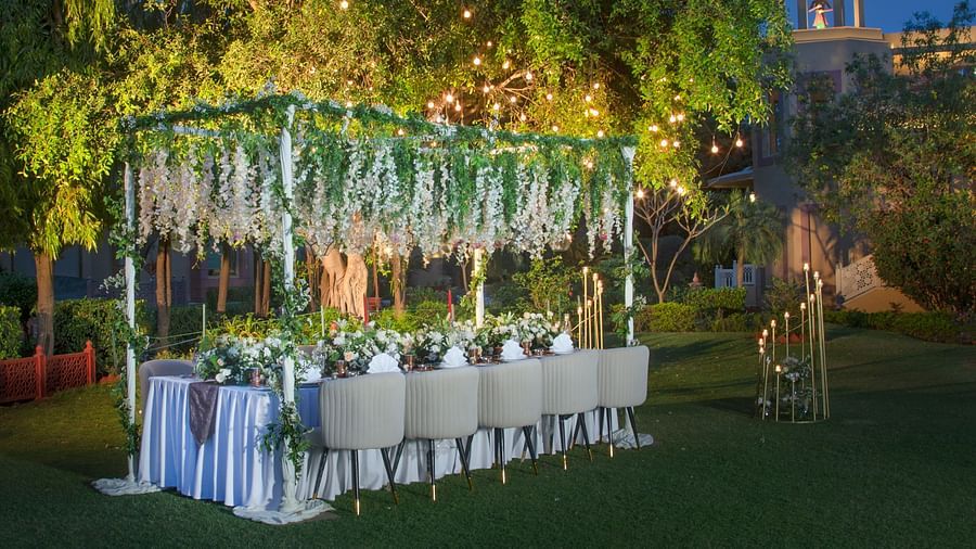 An outdoor evening event with a long dining table set for a meal, adorned in a white tablecloth, surrounded by nature and illuminated by overhead string lights.