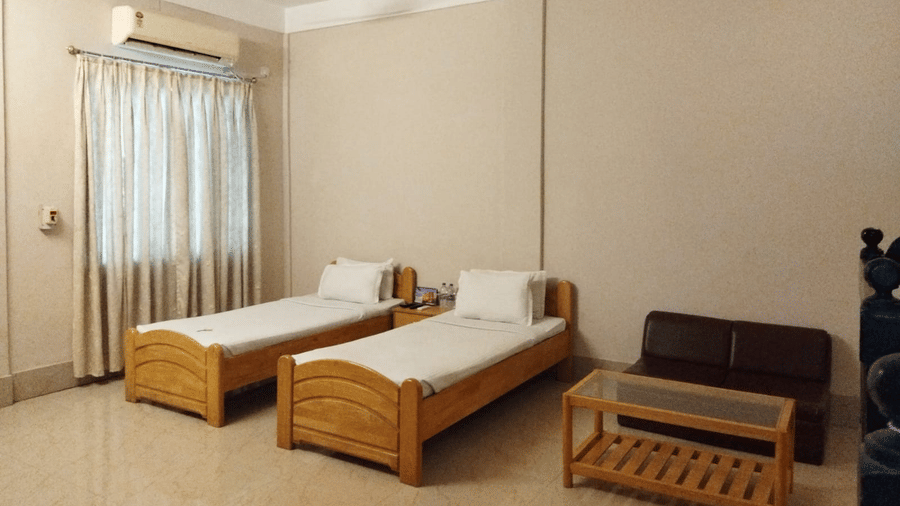Interiors of a room at Lake Side Resort Neermahal featuring 2 wooden beds, an A/C unit, a small wooden table and tiled floors.