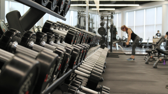 A gym with dumbbells in front and a woman exercising in the back.