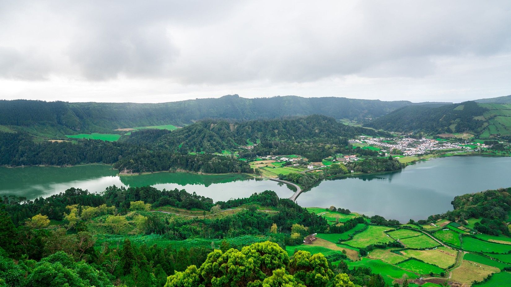A scenic view of a large lake surrounded by rolling hills, agricultural fields, and a small town, with forested areas and cloudy skies in the background.