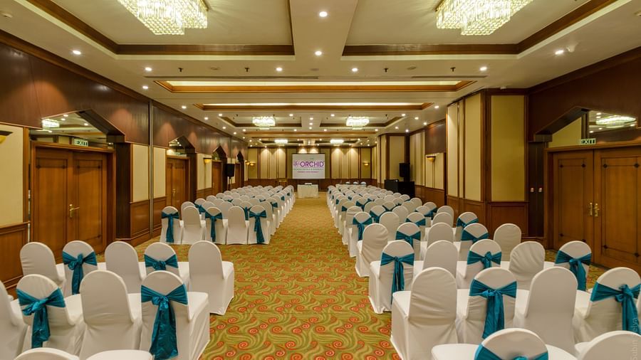 Spacious event hall at The Orchid Hotel arranged with rows of white chairs with teal bows, large chandeliers, and a welcoming atmosphere for large gatherings or conferences.