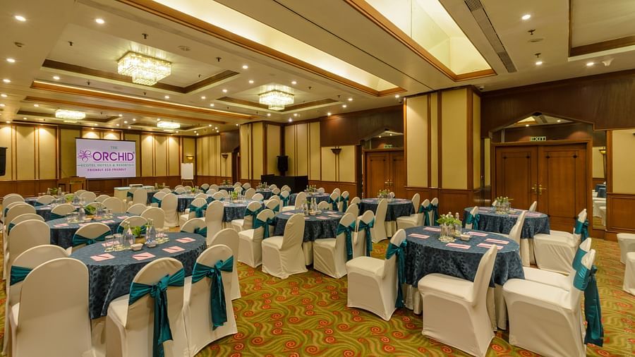 Boardroom meeting setup at The Orchid Hotel, featuring a long table with blue tablecloths and white chairs, overhead chandeliers, and a presentation screen at the front, ideal for corporate meetings.