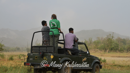 Tourists standing in an open-top safari vehicle while scanning a landscape with distant hills.