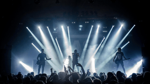 A band performs on a brightly lit stage before a crowd of cheering fans with raised hands.