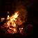 Bright orange flames and smoke rise from a campfire burning wood at night.