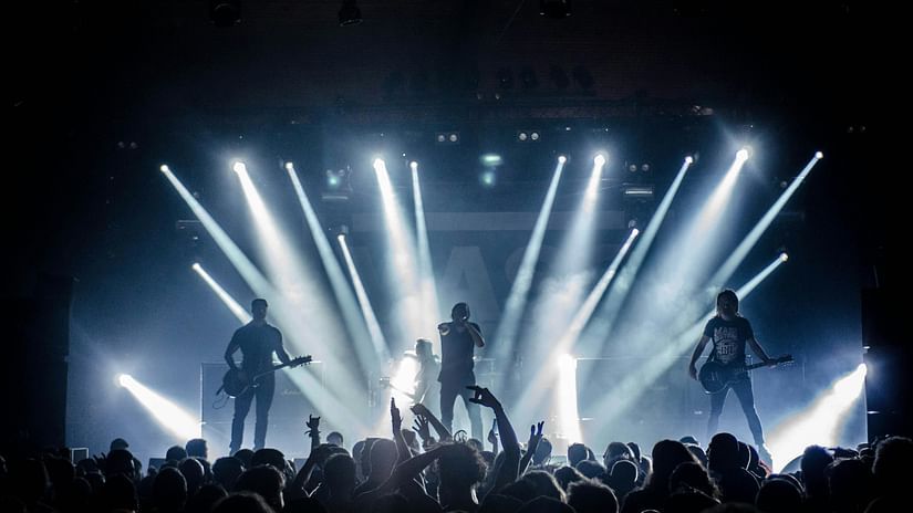 A band performs on a brightly lit stage before a crowd of cheering fans with raised hands.