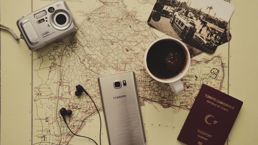 Travel essentials including a camera, passport, smartphone, earphones, and coffee arranged on a vintage city map.