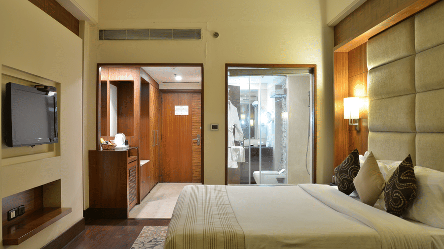 Premium Room at The Bristol Hotel, Gurgaon with a king-size bed, wall-mounted TV, a wooden wardrobe area, and a glass-enclosed bathroom with a walk-in shower.