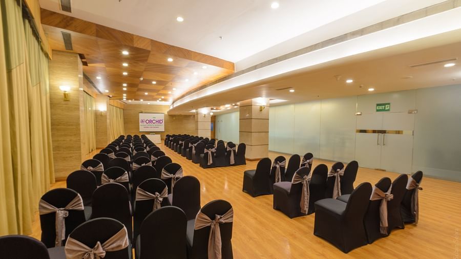 Modern conference room at The Orchid Hotel, featuring rows of black chairs with silver bows, wooden floors, and a projector screen, ready for a professional event.