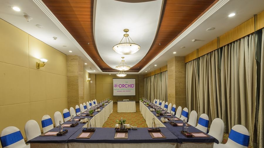 Elegant banquet setup at The Orchid Hotel with round tables dressed in white linen and blue accents, chandeliers overhead, and a vibrant, patterned carpet, creating a festive atmosphere.