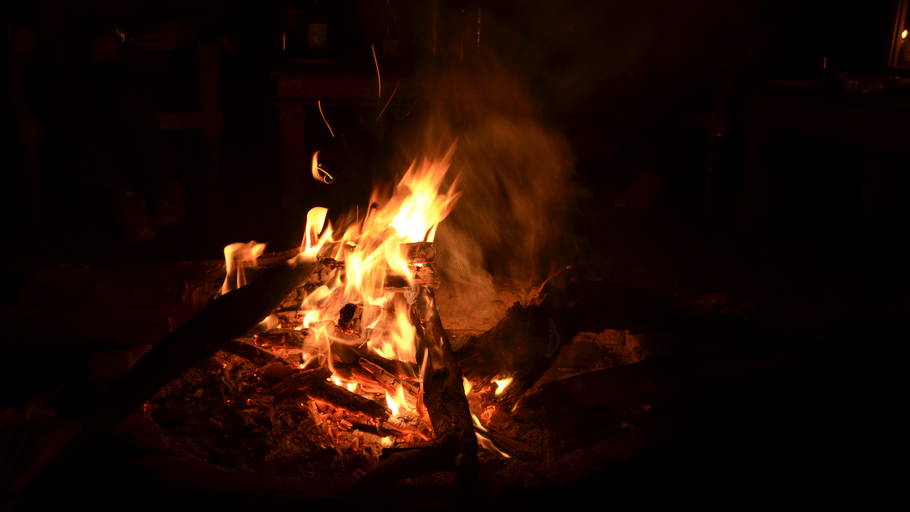 Bright orange flames and smoke rise from a campfire burning wood at night.