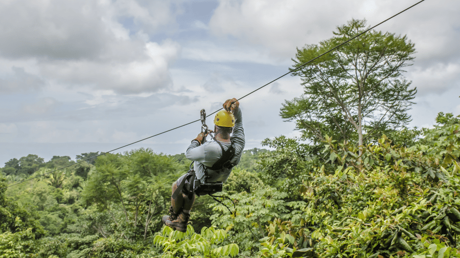 A close-up of a man ziplining through a lush green forest, wearing a helmet and harness, with an excited expression as he glides above the treetops.