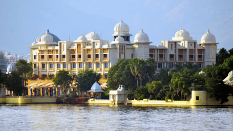 An overview of City Palace in front of Lake Pichola with green trees in front of the building and blue sky in the background. This palace is a must-visit on your 2-day itinerary for Udaipur.