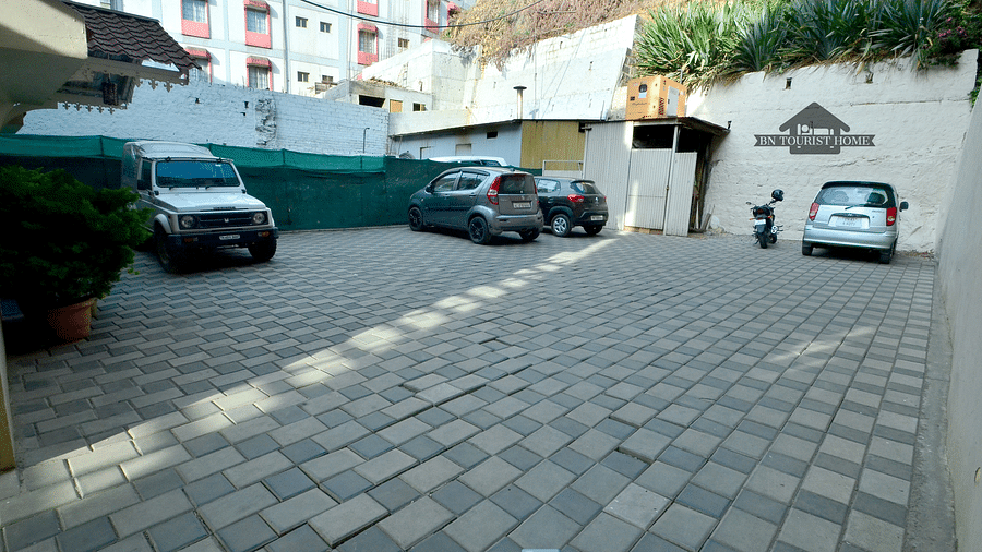 A paved parking lot with several cars and a scooter parked. The lot is surrounded by buildings and greenery -  BN Tourist Home, Ooty