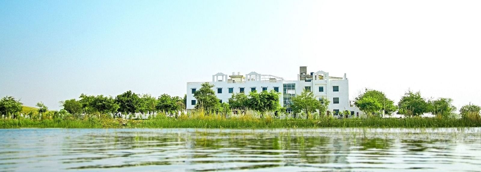 A lake view, with Grassfield Riveria Resort, Jaipur, visible on the shore surrounded by greenery under a clear sky.