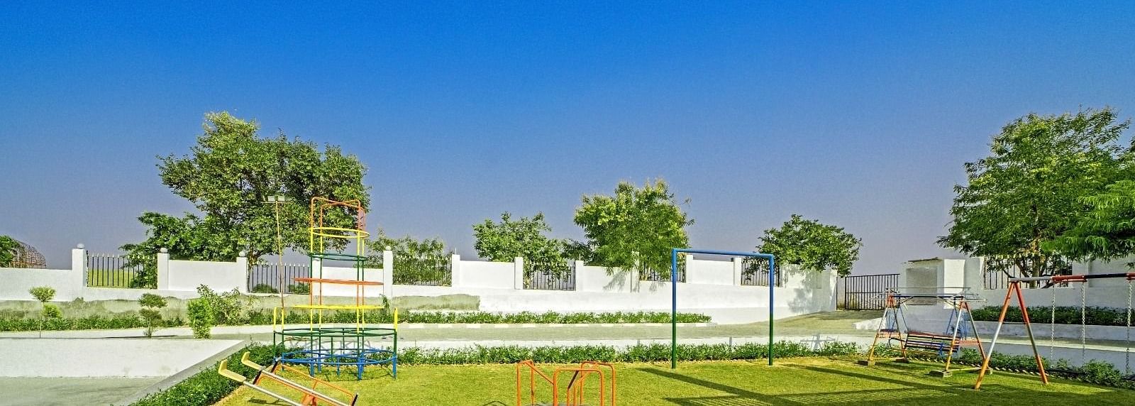 Children’s playground at Grassfield Riveria Resort, Jaipur, featuring swings, a roundabout, and lush greenery under a clear blue sky.