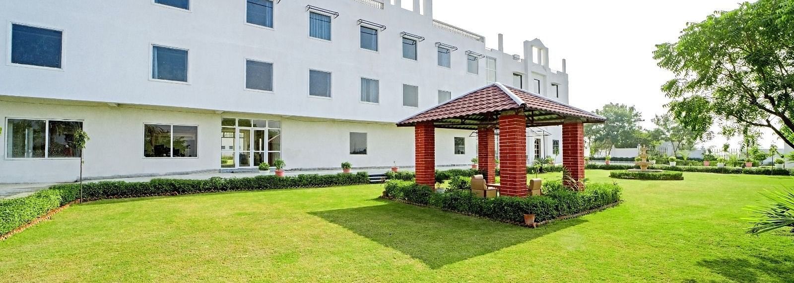 A lawn with a small red gazebo in front of a modern white building at Grassfield Riveria Resort, Jaipur.