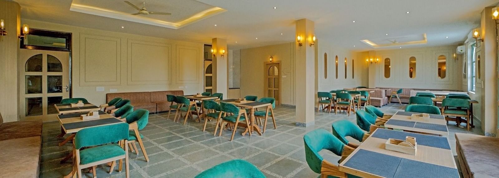Elegant dining area at Grassfield Riveria Resort, Jaipur, with teal upholstered chairs, wooden tables, and warm lighting.