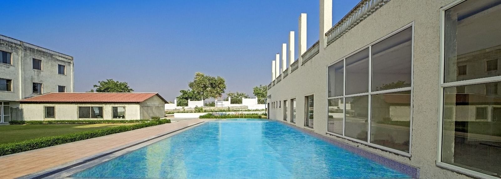 Outdoor swimming pool at Grassfield Riveria Resort, Jaipur, surrounded by modern buildings and a wooden deck area.