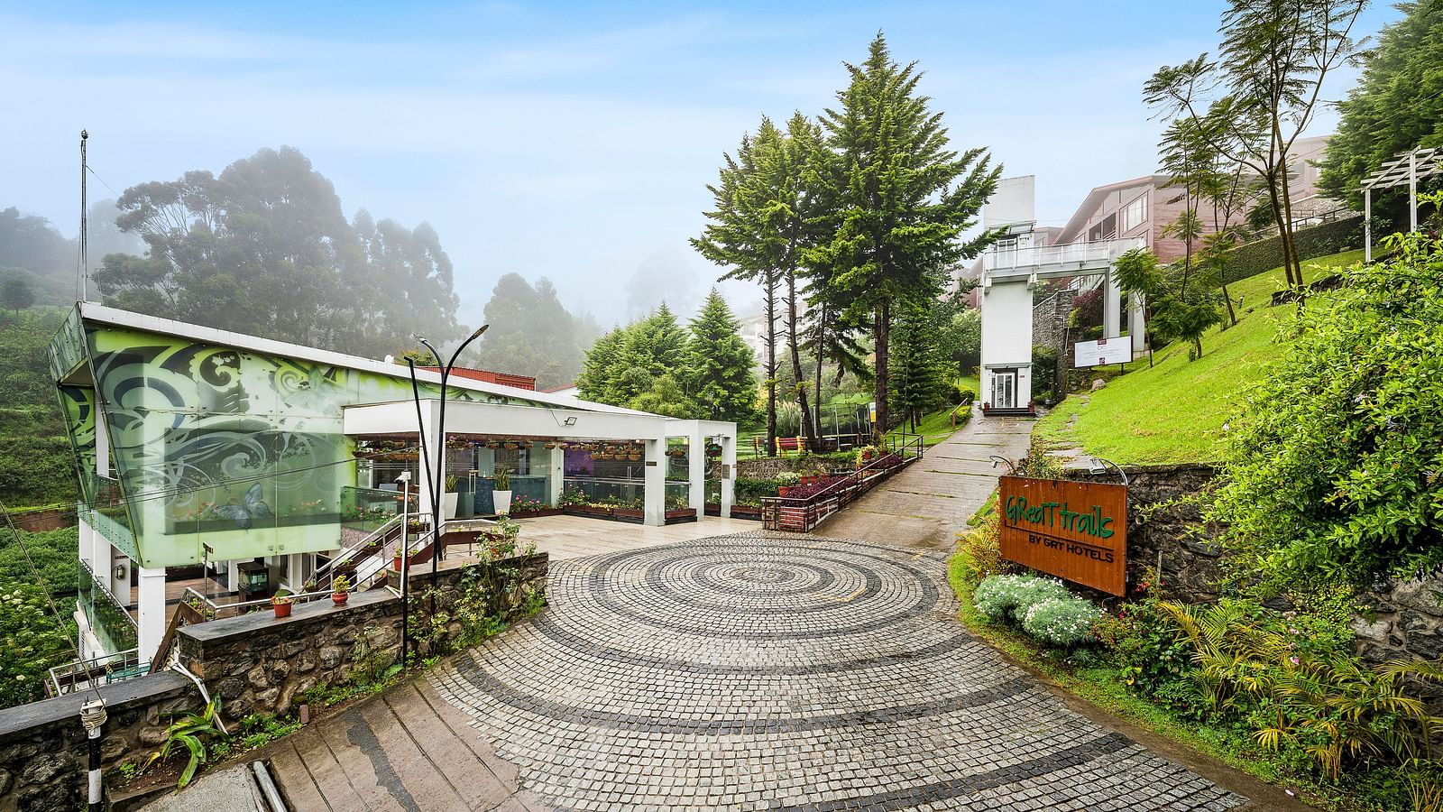 A facade image of the GReaT Trails Kodaikanal By GRT Hotels with lush greenery and the trails leading to the best mountain view resort, Kodaikanal, Tamil Nadu