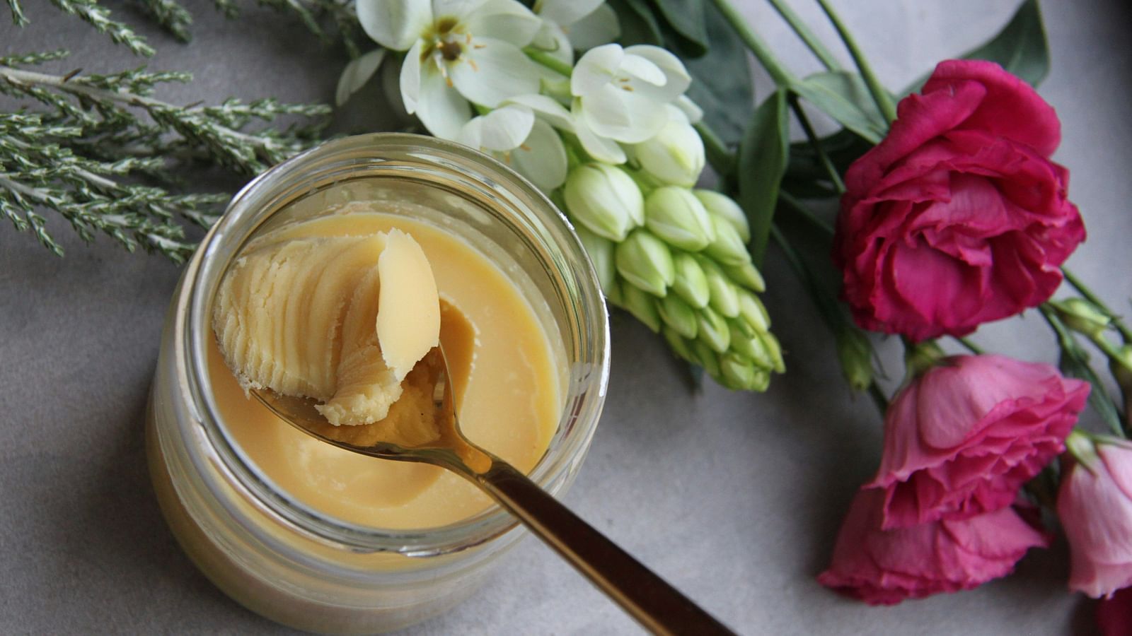 An image featuring a ghee jar with a spoon about to scoop ghee, accompanied by a bunch of flowers placed next to the jar.