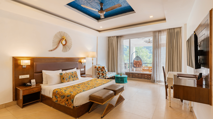 A well-lit bedroom featuring a big bed facing a television screen, with an attached balcony featuring a standing swing chair overlooking the trees outside | Summit by the Ganges Beach Resort & Spa, Rishikesh