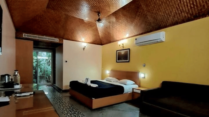 A cosy room with a queen bed, a sofa, wooden furnished ceiling and table with marble floor at Paradise Lagoon Resort, Udupi.